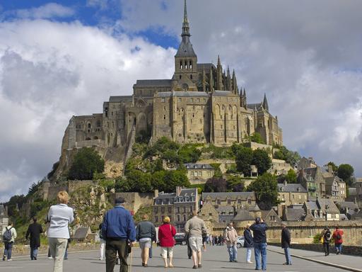 Touristen laufen auf einer Straße auf den Ort Mont-Saint-Michel mit der Abtei in der Normandie in Frankreich zu.