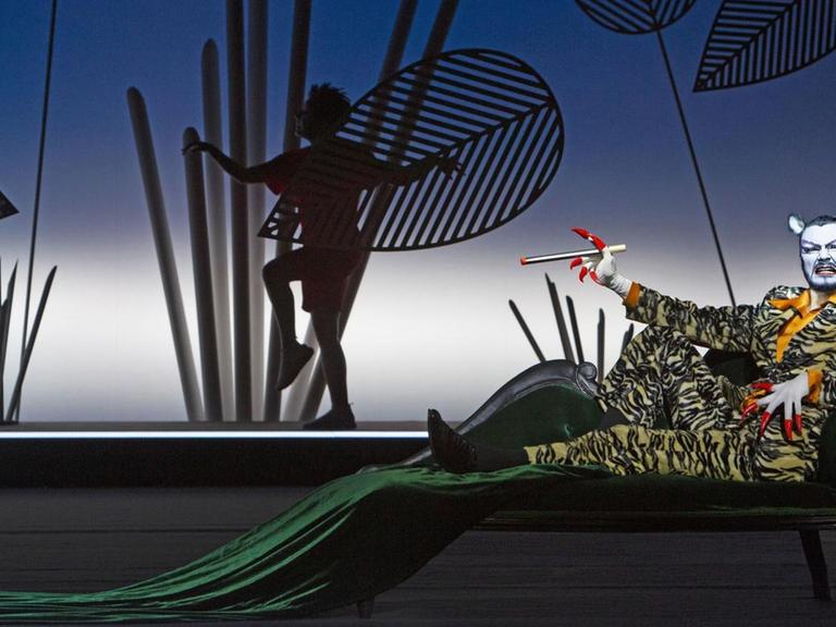 Shere Khan liegt im Vordergrund auf einer Liege, im Hintergrund ist der Schatten von Mowgli sehen sowie Scherenschnitte von Pflanzen.