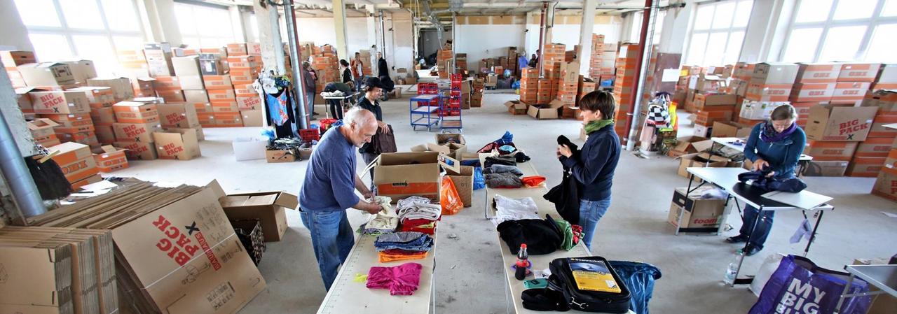 Man sieht eine große Halle mit Kartons voller Kleidung, an Tischen stehen Menschen und sortieren die Spenden. Man sieht eine große Halle mit Kartons voller Kleidung, an Tischen stehen Menschen und sortieren die Spenden.