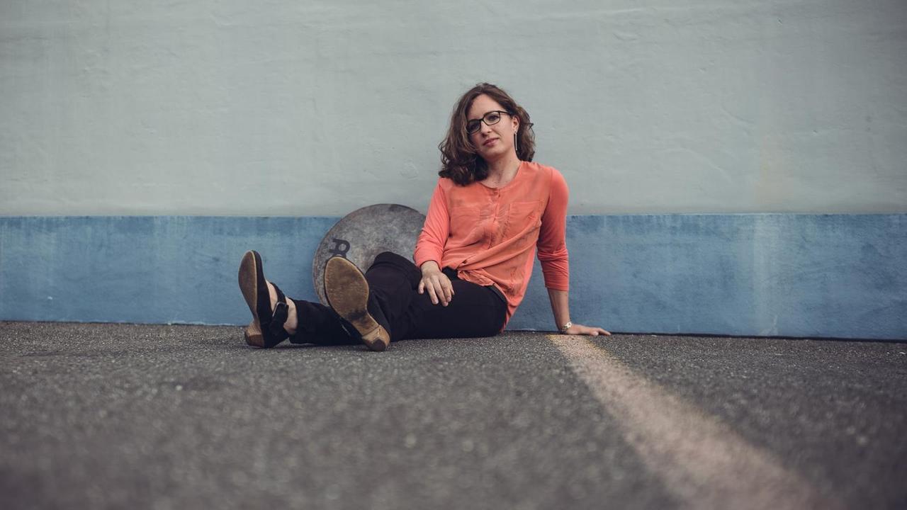 Eine Frau sitzt an eine Wand gelehnt auf dem Boden, neben ihr ist ein einzelnes Schlagzeugbecken aufgestellt