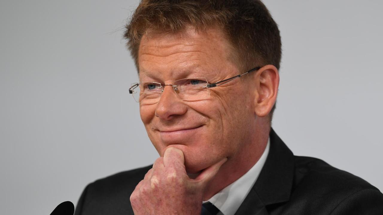 Lutz sitzt vor einem Mikrofon, lächelt und hat eine Hand am Kinn. Er trägt eine Brille. Lutz sitzt vor einem Mikrofon, lächelt und hat eine Hand am Kinn. Er trägt eine Brille.