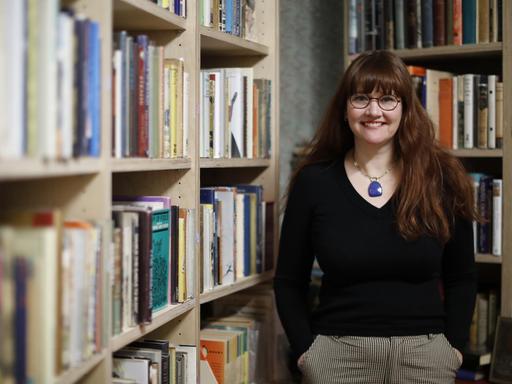 Allison Devers steht in ihrem Buchladen "Second Shelf" in London, umgeben von gefüllten Bücherregalen und lächelt in die Kamera, die Hände in den Hosentaschen.