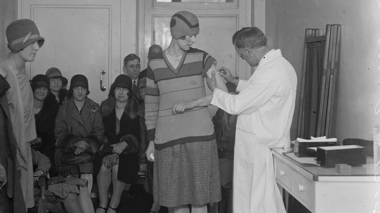 Pockenimpfung im April 1929 in London. Ein Arzt impft in einem Arztzimmer eine Frau. Im Hintergrund warten Frauen und Männer. Pockenimpfung im April 1929 in London. Ein Arzt impft in einem Arztzimmer eine Frau. Im Hintergrund warten Frauen und Männer.