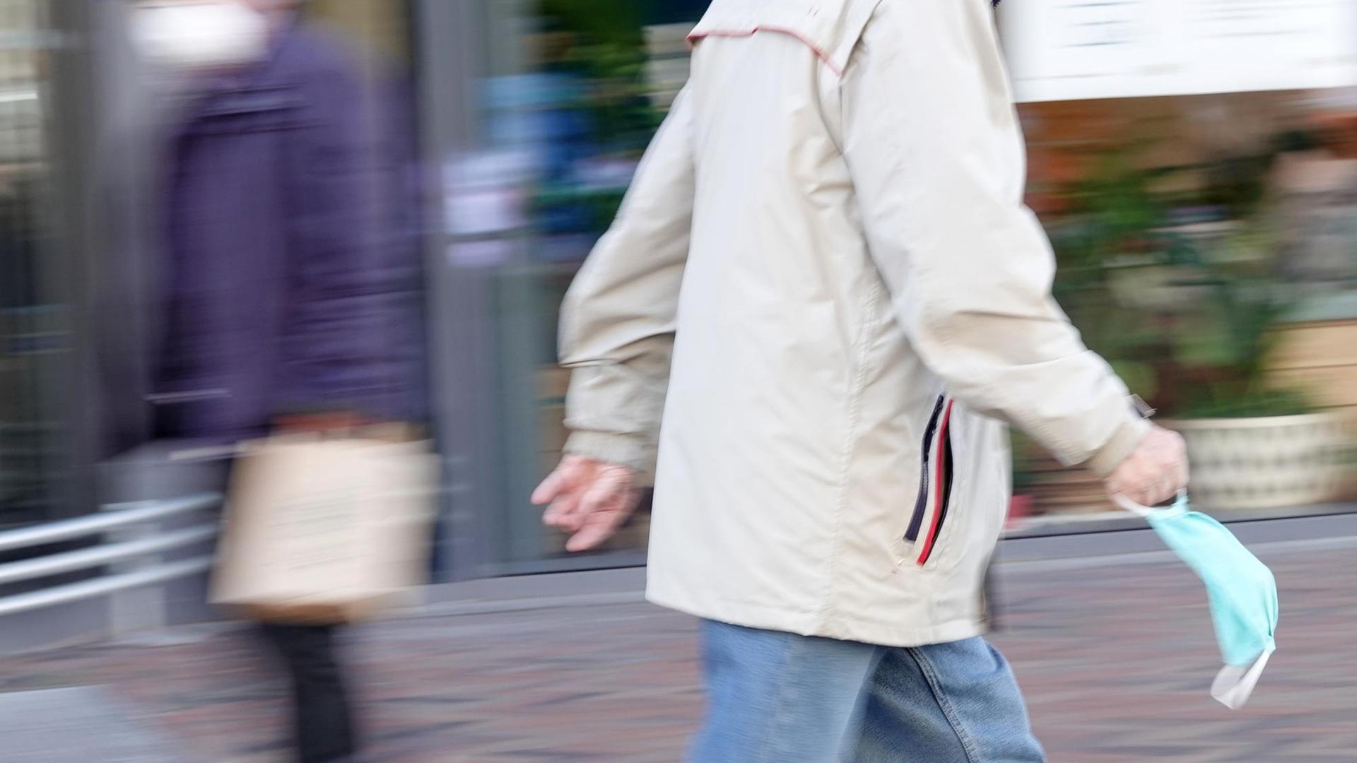 Ein älterer Passant geht durch eine Fußgängerzone und hält eine Mundschutzmaske in der Hand. Im Hintergrund geht weiterer Passant, der einen Mundschutz trägt. Ein älterer Passant geht durch eine Fußgängerzone und hält eine Mundschutzmaske in der Hand. Im Hintergrund geht weiterer Passant, der einen Mundschutz trägt.