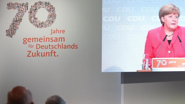Angela Merkel auf einem Videobildschirm im Berliner E-Werk, links daneben der Text "70 Jahre gemeinsam für Deutschlands Zukunft."