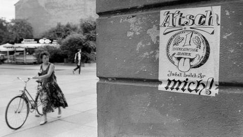 Eine Frau schiebt ein Fahrrad im Bildhintergrund. Im Vordergrund klebt ein Plakat mit einer D-Mark-Karikatur an der Mauer. Es trägt die Aufschrift: "Ätsch, jetzt habt ihr mich!" Darauf zu sehen ist eine D-Mark-Münze, die die Zunge herausstreckt.