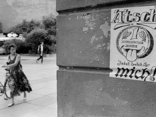 Eine Frau schiebt ein Fahrrad im Bildhintergrund. Im Vordergrund klebt ein Plakat mit einer D-Mark-Karikatur an der Mauer. Es trägt die Aufschrift: "Ätsch, jetzt habt ihr mich!" Darauf zu sehen ist eine D-Mark-Münze, die die Zunge herausstreckt.