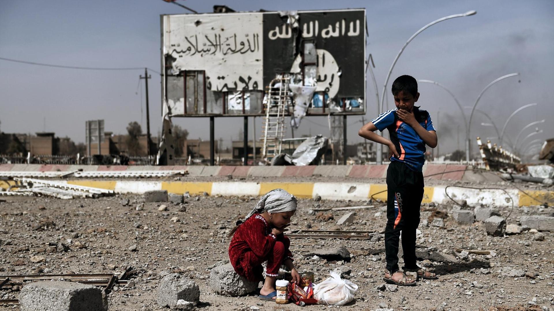 Iraqi children sit amidst the rubble of a street in Mosul's Nablus neighbourhood infront of a billboard bearing the logo of the Islamic State (IS) group on March 12, 2017, during an offensive by security forces to retake the western parts of the city from IS fighters. / AFP PHOTO / ARIS MESSINIS Iraqi children sit amidst the rubble of a street in Mosul's Nablus neighbourhood infront of a billboard bearing the logo of the Islamic State (IS) group on March 12, 2017, during an offensive by security forces to retake the western parts of the city from IS fighters. / AFP PHOTO / ARIS MESSINIS