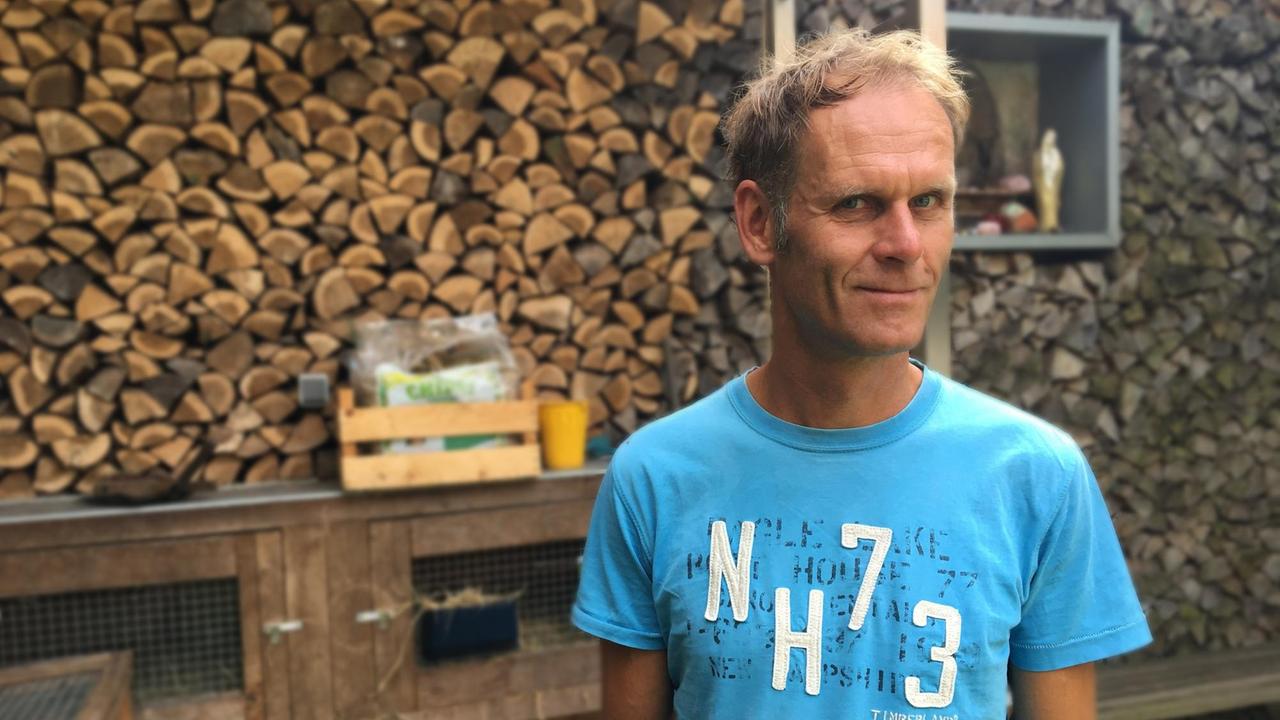 Der Baumhausarchitekt Andreas Wenning steht in einem blauen T-Shirt vor einem Stapel Feuerholz.
