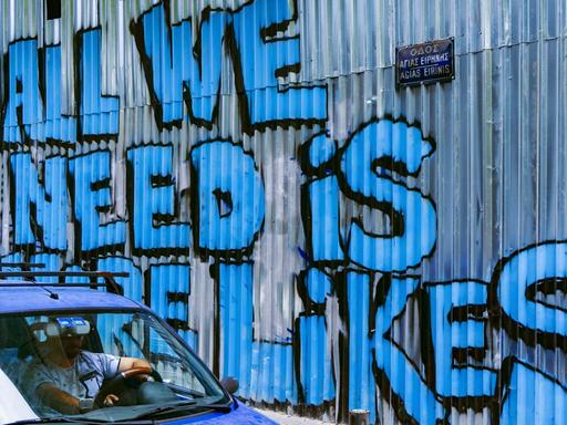 Ein Auto passiert eine Wellblechwand mit einem Graffiti, das in großer blauer Schrift verkündet: "All We Need Is More Likes".