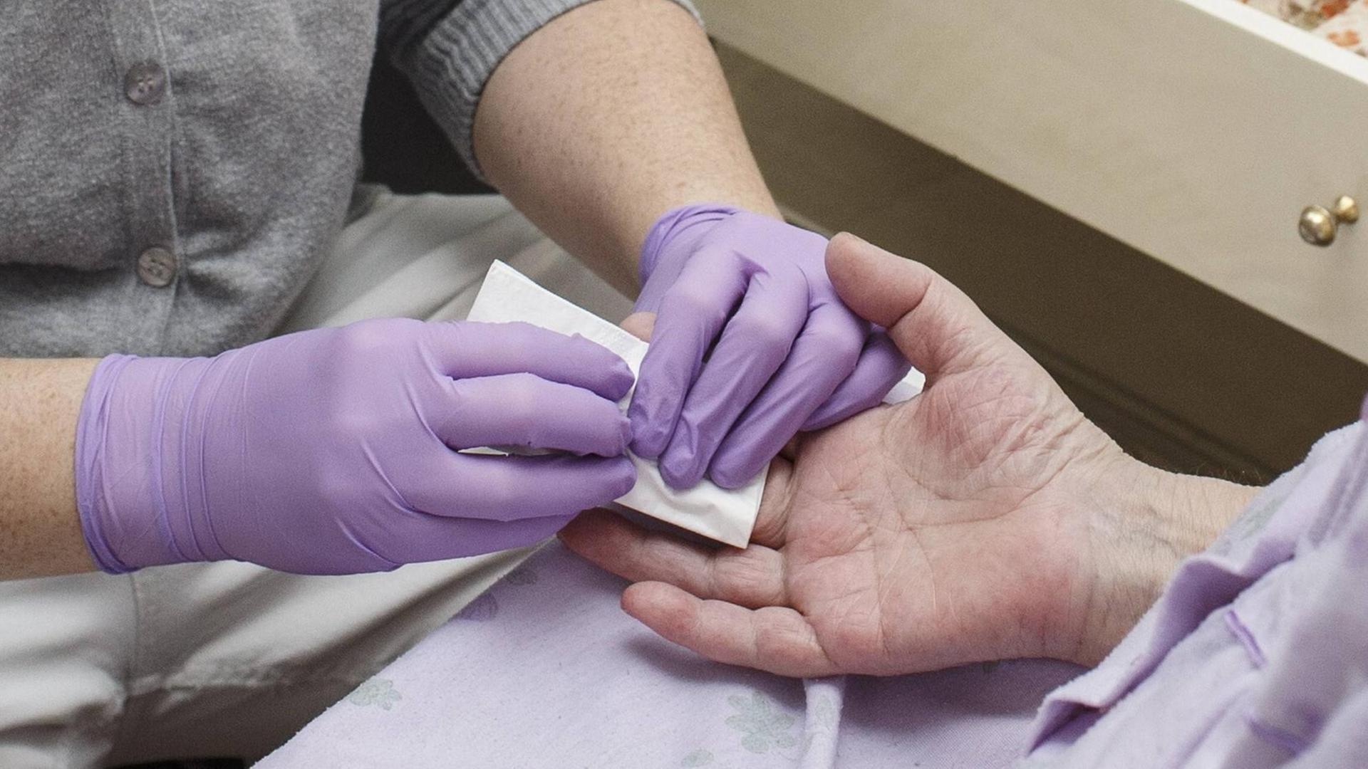 Eine Mitarbeiterin eines Pflegedienstes misst einer Patienten den Blutzuckerwert. Mit lilafarbenen Gummihandschuhen tupft sie einen Bluttropfen v on der Hand der Patientin ab. Eine Mitarbeiterin eines Pflegedienstes misst einer Patienten den Blutzuckerwert. Mit lilafarbenen Gummihandschuhen tupft sie einen Bluttropfen v on der Hand der Patientin ab.