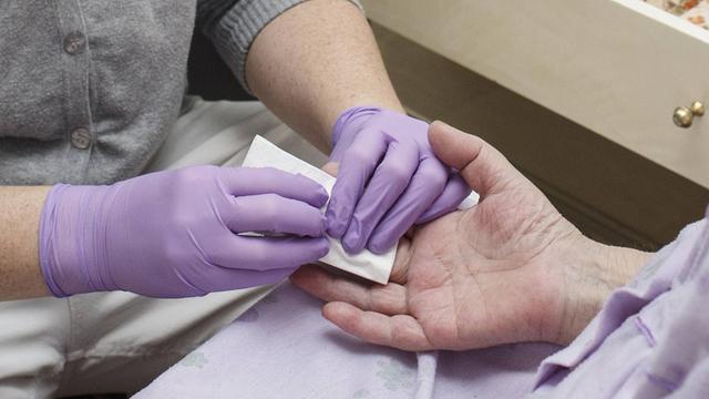 Eine Mitarbeiterin eines Pflegedienstes misst einer Patienten den Blutzuckerwert. Mit lilafarbenen Gummihandschuhen tupft sie einen Bluttropfen v on der Hand der Patientin ab.
