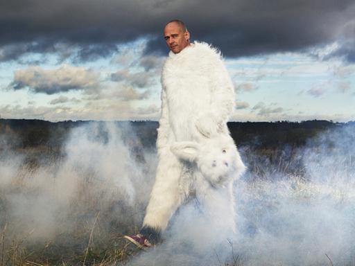 Das Bild zeigt den britischen Künstler Dinos Chapman in weißem Hasenkostüm. Den Hasenkopf in der Hand haltend, geht er durch eine neblige Landschaft.