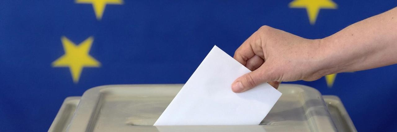 Eine Hand steckt einen Umschlag in eine Wahlurne vor der Europafahne. Symbolfoto. Eine Hand steckt einen Umschlag in eine Wahlurne vor der Europafahne. Symbolfoto.