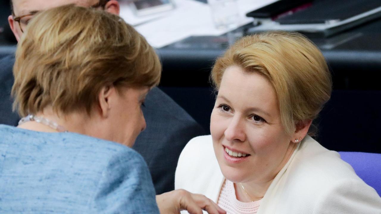 Giffey sitzt auf ihrem Stuhl und hört lächelnd Merkel zu, die sich zu ihr vorbeugt und redet. Merkel ist unscharf im Vordergrund zu sehen.