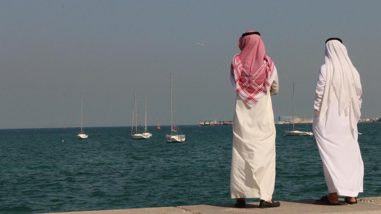 Zwei traditionell gekleidete katarische Bürger stehen an der Uferpromenade der Hauptstadt Doha und blicken auf den Persischen Golf. Zwei traditionell gekleidete katarische Bürger stehen an der Uferpromenade der Hauptstadt Doha und blicken auf den Persischen Golf.