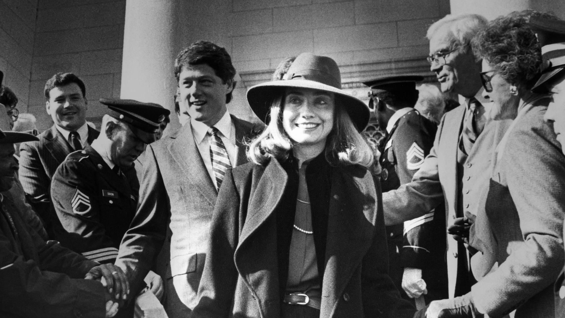 Historisches Foto von Hillary Clinton im Wahlkampf von ihrem Mann, in einer Menschenmenge beim Hände schütteln, im Hintergrund Bill Clinton.