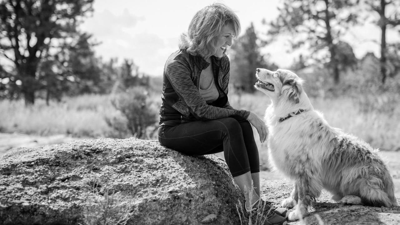 Eine Frau sitzt auf einem Stein und lächelt ihren Hund an, der zu ihr aufschaut. Eine Frau sitzt auf einem Stein und lächelt ihren Hund an, der zu ihr aufschaut.