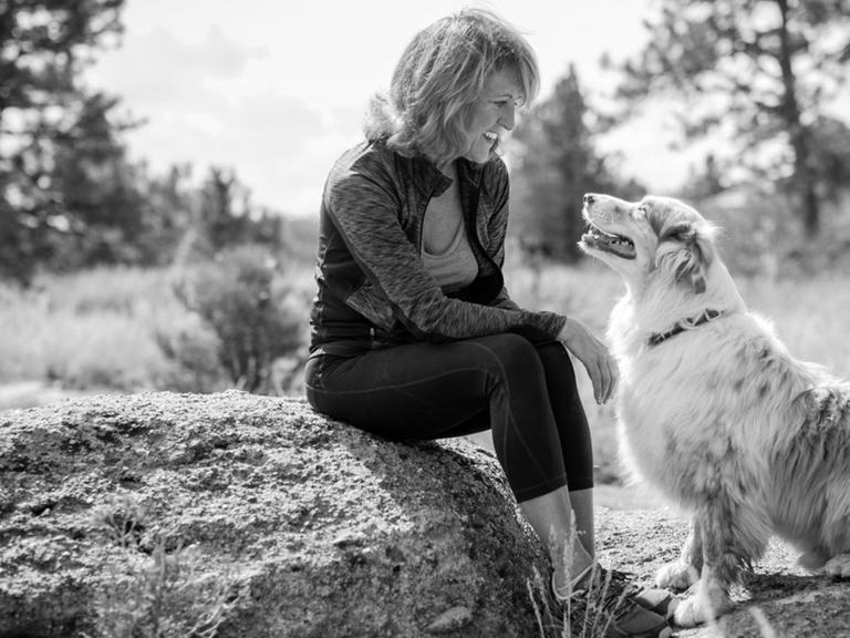 Eine Frau sitzt auf einem Stein und lächelt ihren Hund an, der zu ihr aufschaut.