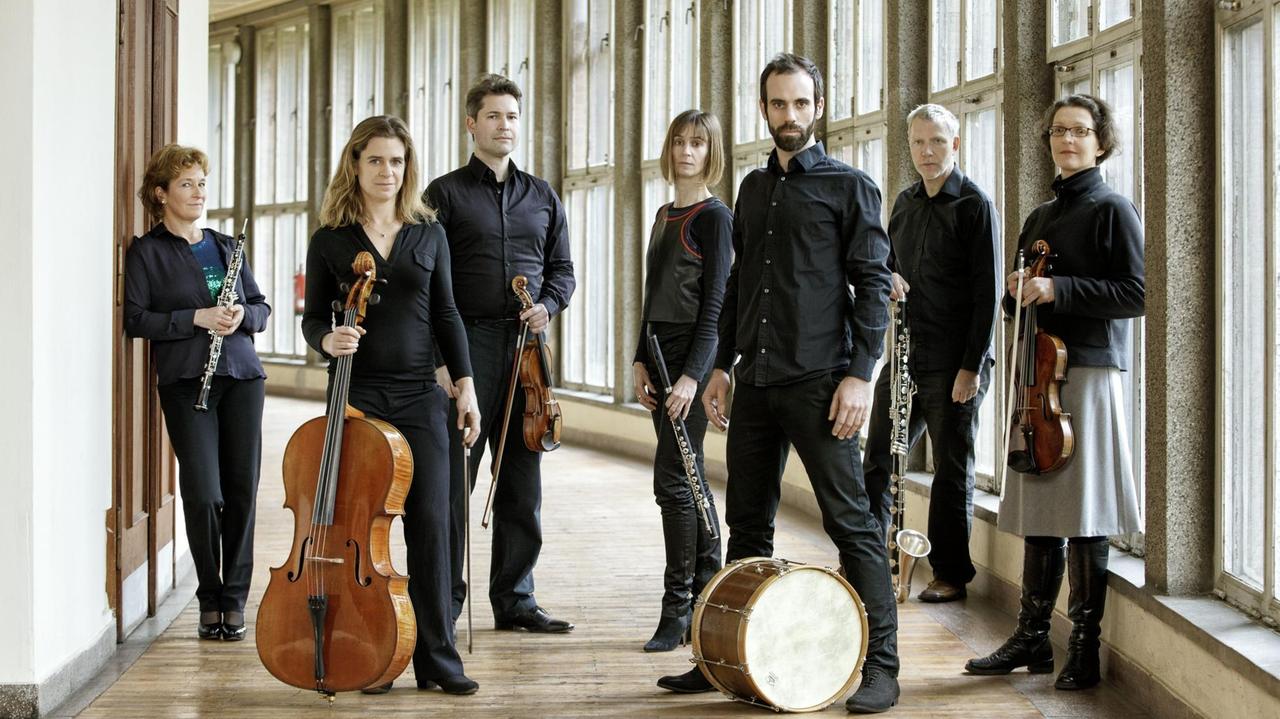 Sieben Musiker stehen mit ihren Instrumenten in einem Gang mit großen Fenstern. Sieben Musiker stehen mit ihren Instrumenten in einem Gang mit großen Fenstern.