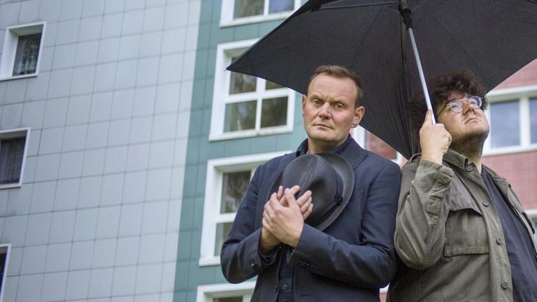 Devid Striesow und Axel Ranisch mit einem schwarzen, quadratischen Regenschirm vor einem Blattenbau.
