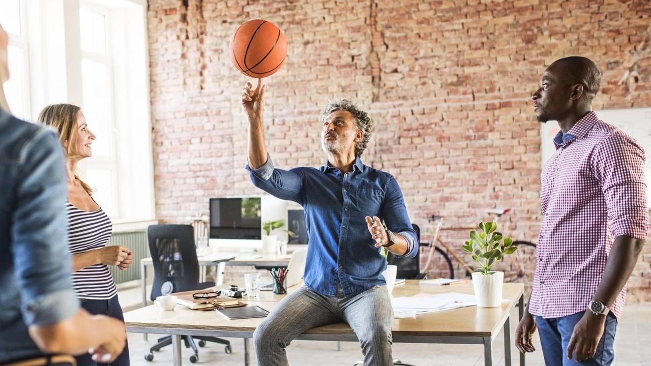 Ein Mann sitzt auf einem Schreibtisch und balanciert einen Basketball auf dem Finger. Um ihn herum stehen Kollegen. Das Büro ist licht, stilvoll und geräumig eingerichtet. Ein Mann sitzt auf einem Schreibtisch und balanciert einen Basketball auf dem Finger. Um ihn herum stehen Kollegen. Das Büro ist licht, stilvoll und geräumig eingerichtet.
