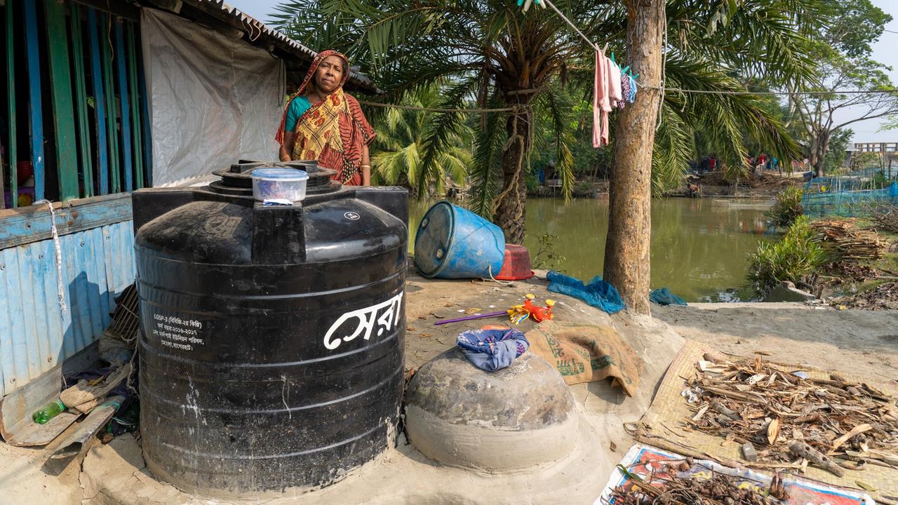 
Purnima Nath steht neben ihrem Haus hinter einem schwarzen Tank mit Süßwasser