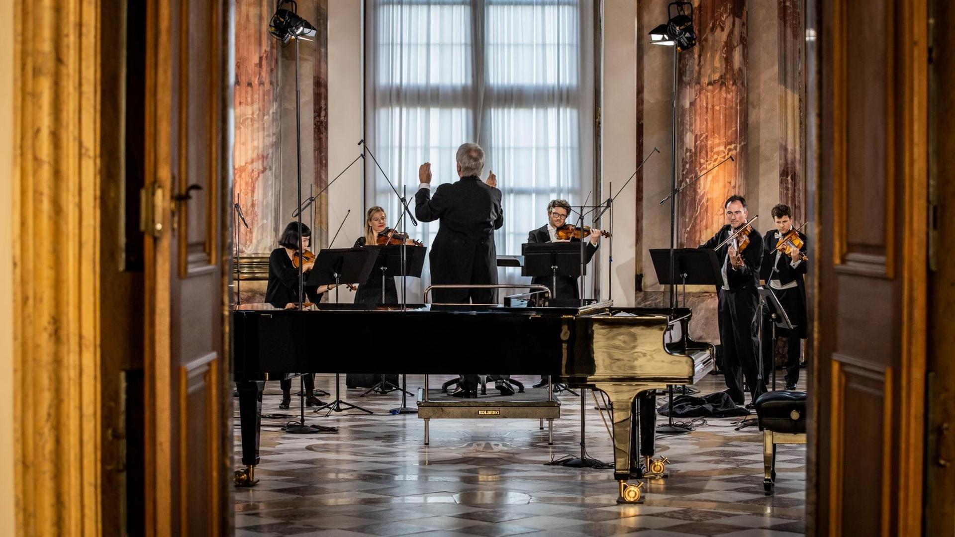 Durch eine geöffnete Tür blickt man auf ein Orchester in einem barocken Saal, das von einem Mann dirigiert wird. Durch eine geöffnete Tür blickt man auf ein Orchester in einem barocken Saal, das von einem Mann dirigiert wird.