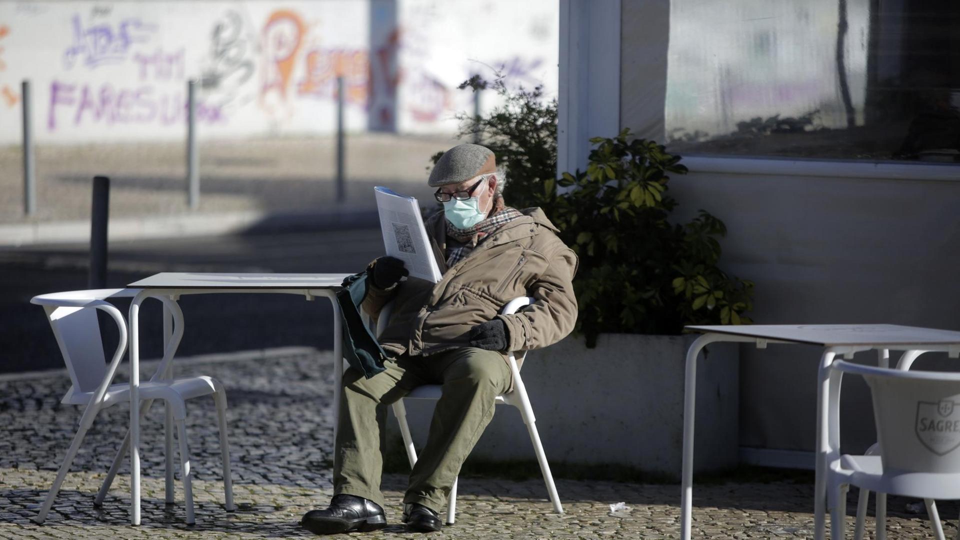 Ein Mann mit einer Maske sitzt alleine an einem leeren Cafétisch in Lissabon und liest Zeitung.