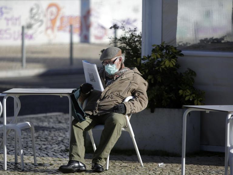 Ein Mann mit einer Maske sitzt alleine an einem leeren Cafétisch in Lissabon und liest Zeitung.