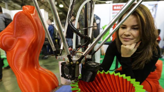 Ein 3D-Drucker entwirft eine rot-grüne Tülle, im Hintergrund schaut eine Frau zu.