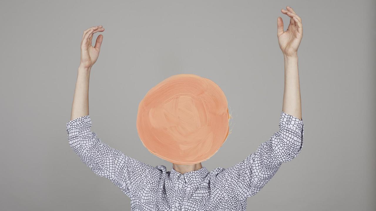 Ein Mensch in gemustertem weißem Hemd hebt die Arme empor – der Kopf ist mit einem orangene Kreis übermalt. Ein Mensch in gemustertem weißem Hemd hebt die Arme empor – der Kopf ist mit einem orangene Kreis übermalt.