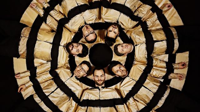 Sieben in schwarz-goldene Hemden gekleidete Männer liegen mit dem Kopf zur Kreismitte auf dem Boden.