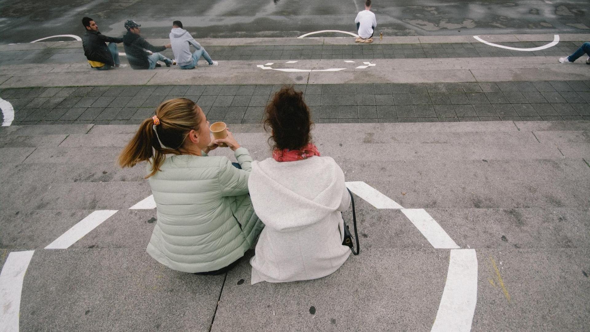 Zwei junge Frauen sitzen mit einem Kaffe auf den Treppenstufen und innerhalb eines eingezeichneten Kreises, um den nötigen Abstand einzuhalten. Zwei junge Frauen sitzen mit einem Kaffe auf den Treppenstufen und innerhalb eines eingezeichneten Kreises, um den nötigen Abstand einzuhalten.