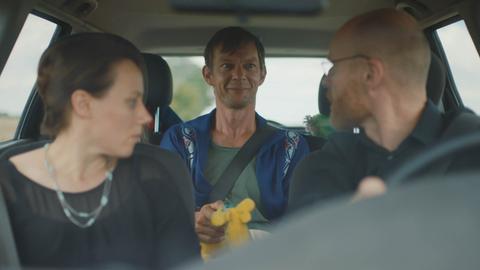 Filmstill aus "Whatever Happens Next" von Regisseur Julian Pörksen: die Hauptfigur Paul sitzt auf dem Rücksitz eines Autos, auf dem Fahrer- und Beifahrersitz ist ein Paar zu sehen
