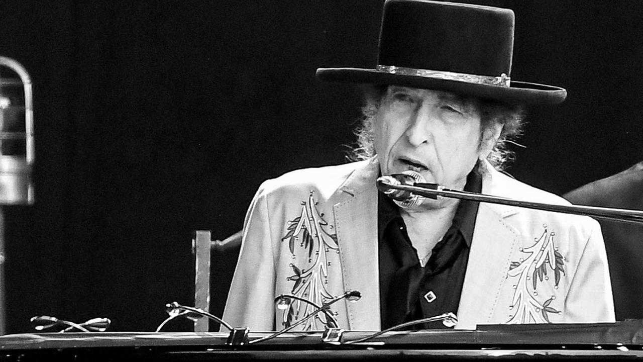 Schwarz-weiß Fotografie von Bob Dylan während eines Auftritts im Hyde Park im vergangenen Jahr. Er sitzt am Klavier, singt ins Mikrofon und trägt einen Hut mit breiter Krempe und Silberband, schwarzes Hemd und hellgrauen Sakko mit Pailletten und Stickmuster.
