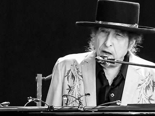 Schwarz-weiß Fotografie von Bob Dylan während eines Auftritts im Hyde Park im vergangenen Jahr. Er sitzt am Klavier, singt ins Mikrofon und trägt einen Hut mit breiter Krempe und Silberband, schwarzes Hemd und hellgrauen Sakko mit Pailletten und Stickmuster.