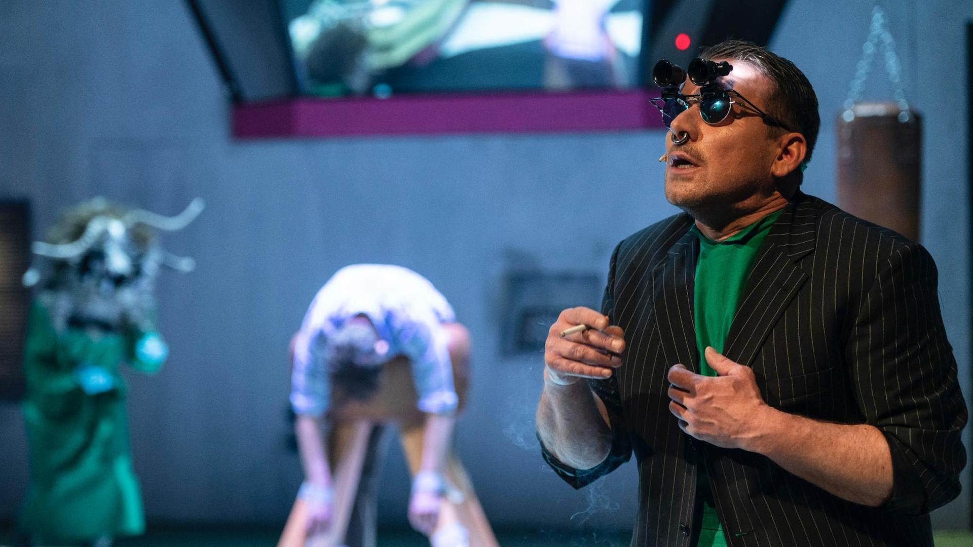 Bühnenszenario: Im Vordergrund steht ein Schauspieler mit Zigarette in der Hand. Er trägt Sonnenbrille mit einem aufgesteckten Fernglas. Im Hintergrund in der Unschärfe weitere Schauspieler.