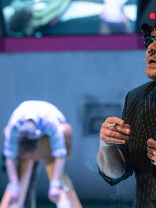 Bühnenszenario: Im Vordergrund steht ein Schauspieler mit Zigarette in der Hand. Er trägt Sonnenbrille mit einem aufgesteckten Fernglas. Im Hintergrund in der Unschärfe weitere Schauspieler.