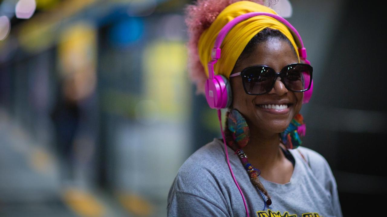 Eine Frau steht vor einem Zug und hört sichtlich glücklich Musik über Kopfhörer.