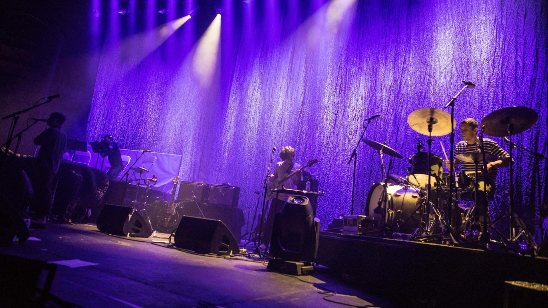Eine Band steht auf der Bühne. Die Beleuchtung ist in blau gehüllt. Zu sehen ist unter anderem ein Schlagzeug. Eine Band steht auf der Bühne. Die Beleuchtung ist in blau gehüllt. Zu sehen ist unter anderem ein Schlagzeug.