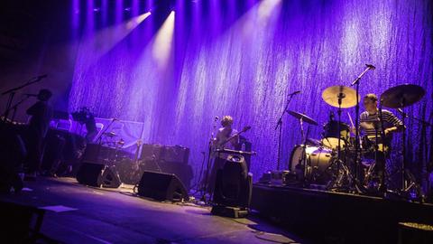 Eine Band steht auf der Bühne. Die Beleuchtung ist in blau gehüllt. Zu sehen ist unter anderem ein Schlagzeug.