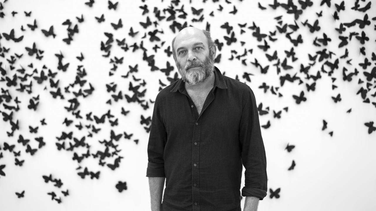 Carlos Amorales steht vor einer weißen Wand, die mit schwarzen Papier-Schmetterlingen beklebt ist.