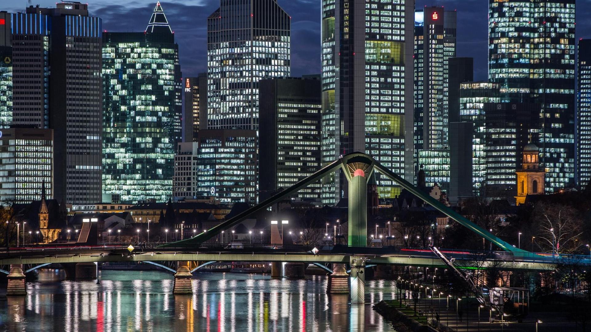 Die hell erleuchteten Büros der Bankentürme hinter einer Brücke über den Main in der Dunkelheit. Die hell erleuchteten Büros der Bankentürme hinter einer Brücke über den Main in der Dunkelheit.