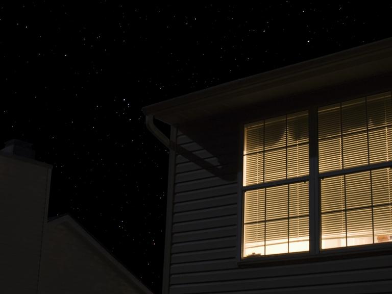 In der Fassade eines Wohnhauses dringt durch ein Fenster mit geschlossenen Jalousien Licht nach außen.
