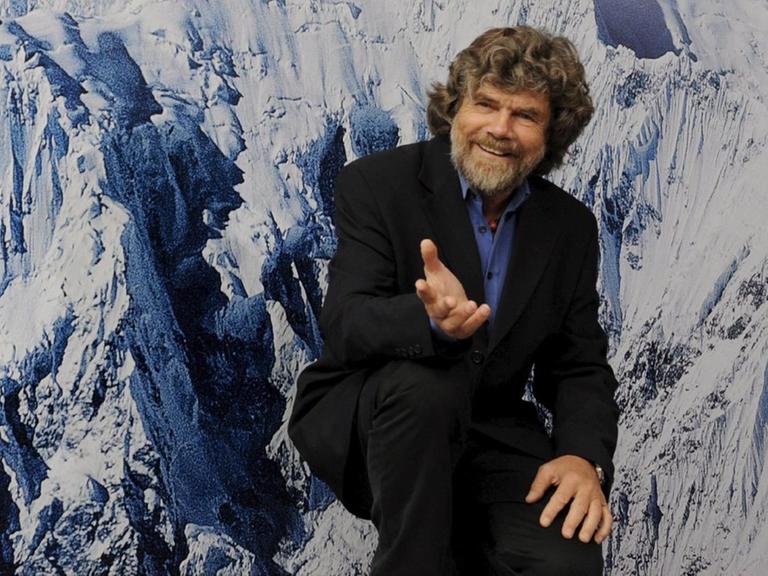 Reinhold Messner sitzt vor einer Fototapete mit schneebedeckten Bergen.