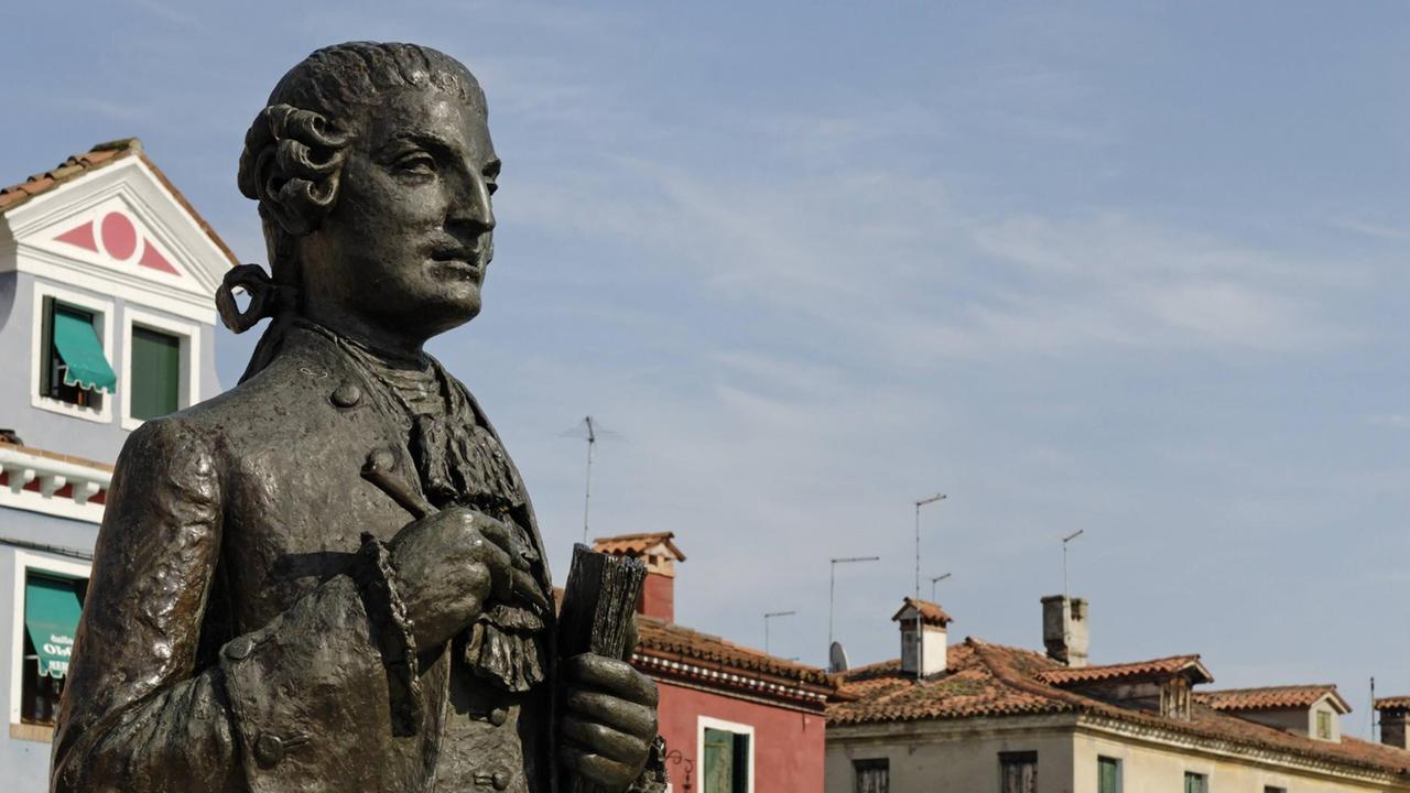 Die Statue von Baldassare Galuppi, die vor den bunten Häusern von Burano steht, hält einen Stift und ein Buch in den Händen. Die Statue von Baldassare Galuppi, die vor den bunten Häusern von Burano steht, hält einen Stift und ein Buch in den Händen.