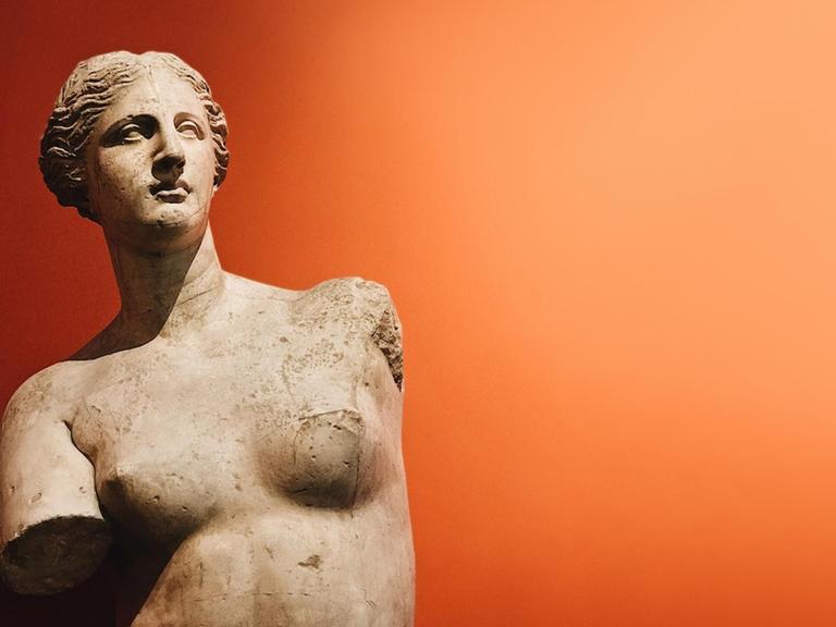 Der Torso einer antiken Frauenstatue vor einem orangefarbenen Hintergrund.
