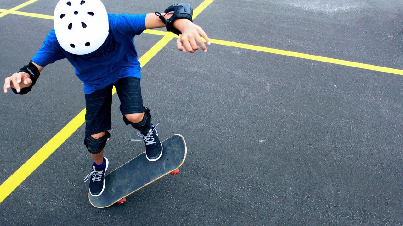 Ein Kind mit Helm und Schonern übt auf einer asphaltierten Fläche mit gelben Gitternetzlinien einen Trick auf dem Skateboard. Ein Kind mit Helm und Schonern übt auf einer asphaltierten Fläche mit gelben Gitternetzlinien einen Trick auf dem Skateboard.