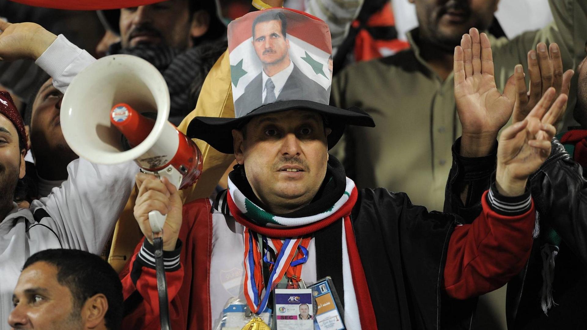 Ein syrischer Fußballfan mit Megaphon trägt bei einem Spiel des Asiencups 2011 das Konterfei von Präsident Baschar al Assad auf dem Hut. Ein syrischer Fußballfan mit Megaphon trägt bei einem Spiel des Asiencups 2011 das Konterfei von Präsident Baschar al Assad auf dem Hut.
